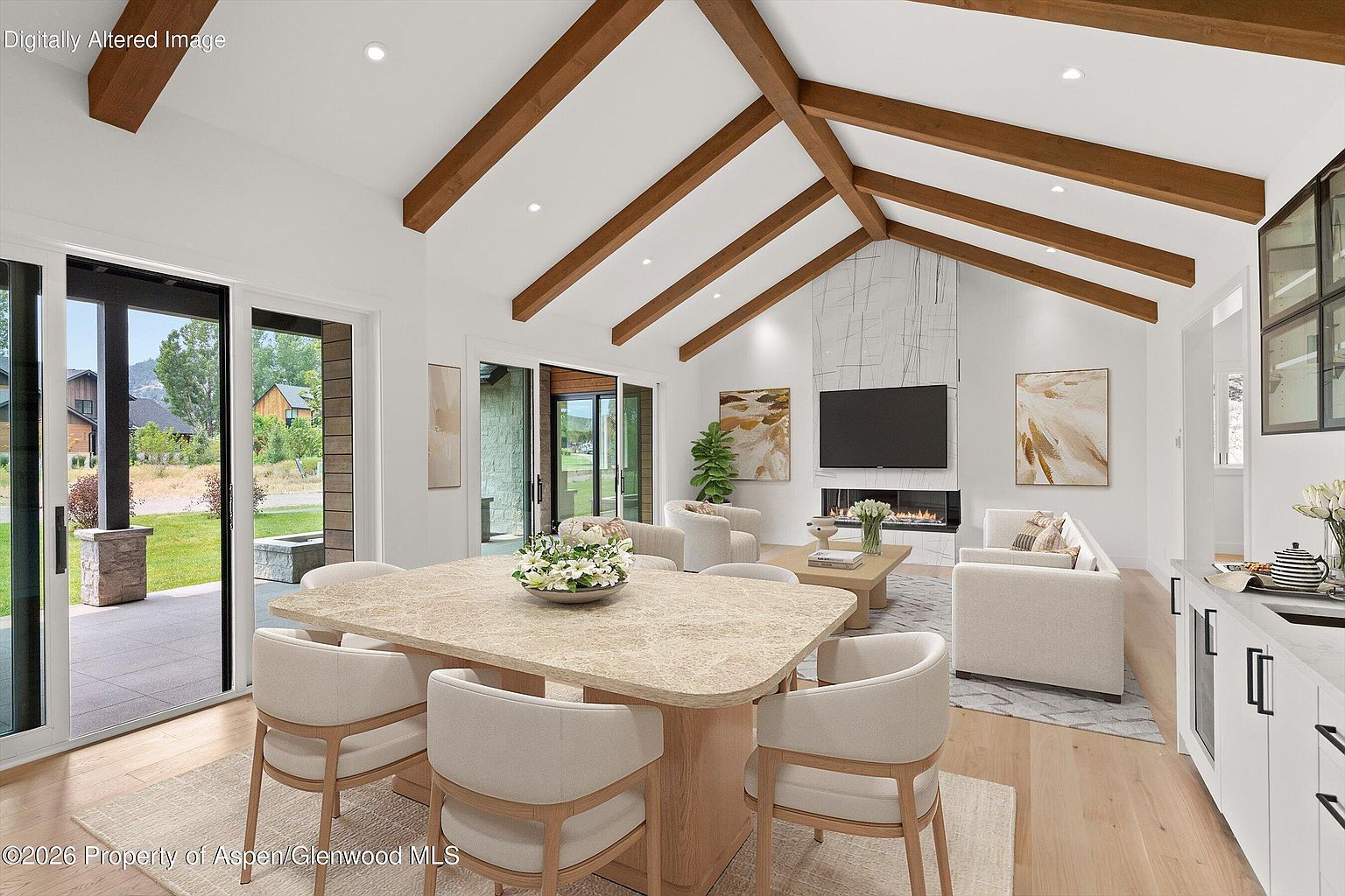 This open-concept living and dining area features vaulted ceilings with exposed wood beams and light-toned wood flooring. A central fireplace with a tall, patterned stone surround serves as a focal point, flanked by two pieces of abstract wall art. Large sliding glass doors provide seamless access to the outdoor patio, creating a bright and airy atmosphere with a view of the surrounding landscape.
