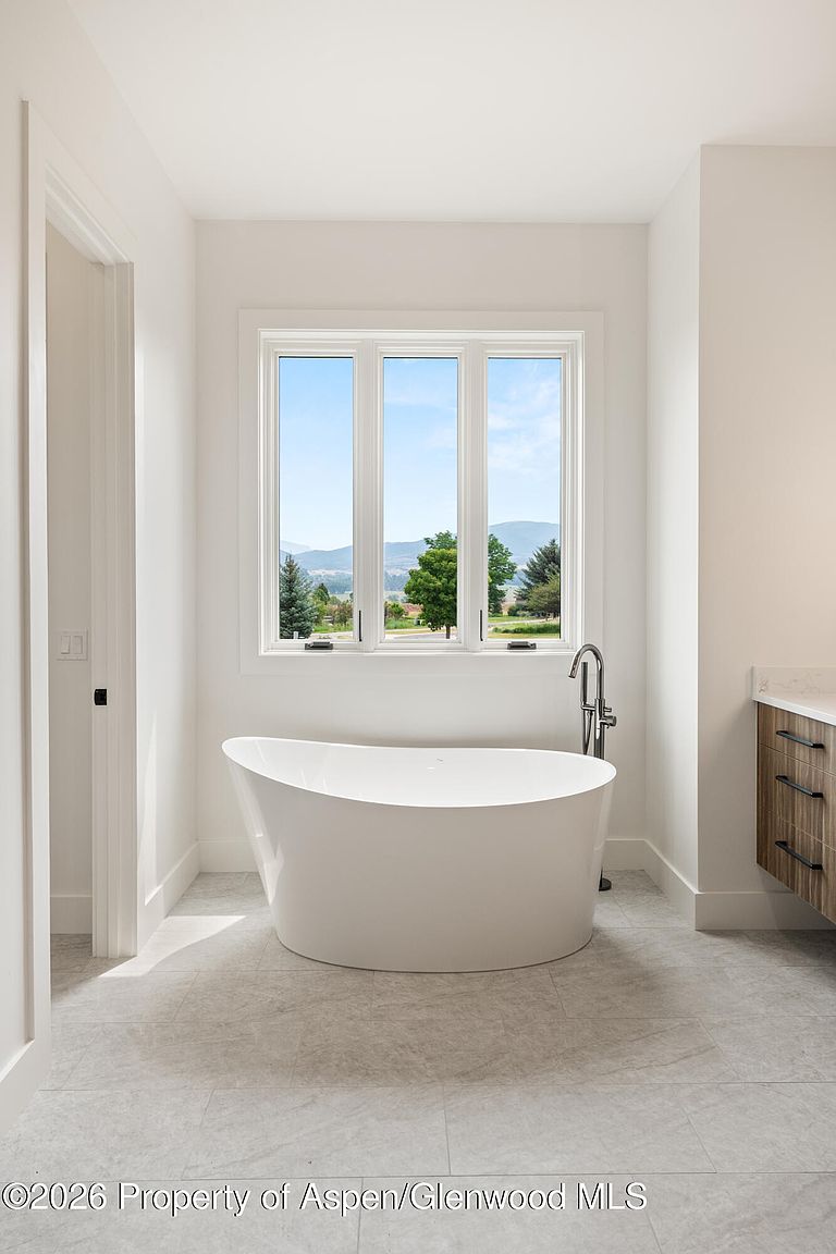 This primary bathroom features a sleek, freestanding soaking tub positioned centrally beneath a large three-pane window that frames a scenic mountain view. To the right, a floating vanity with wood-toned cabinetry and dark hardware adds a modern touch, while the room is finished with neutral, large-format floor tiles. The space is bright and minimalist, emphasizing a clean, spa-like atmosphere.