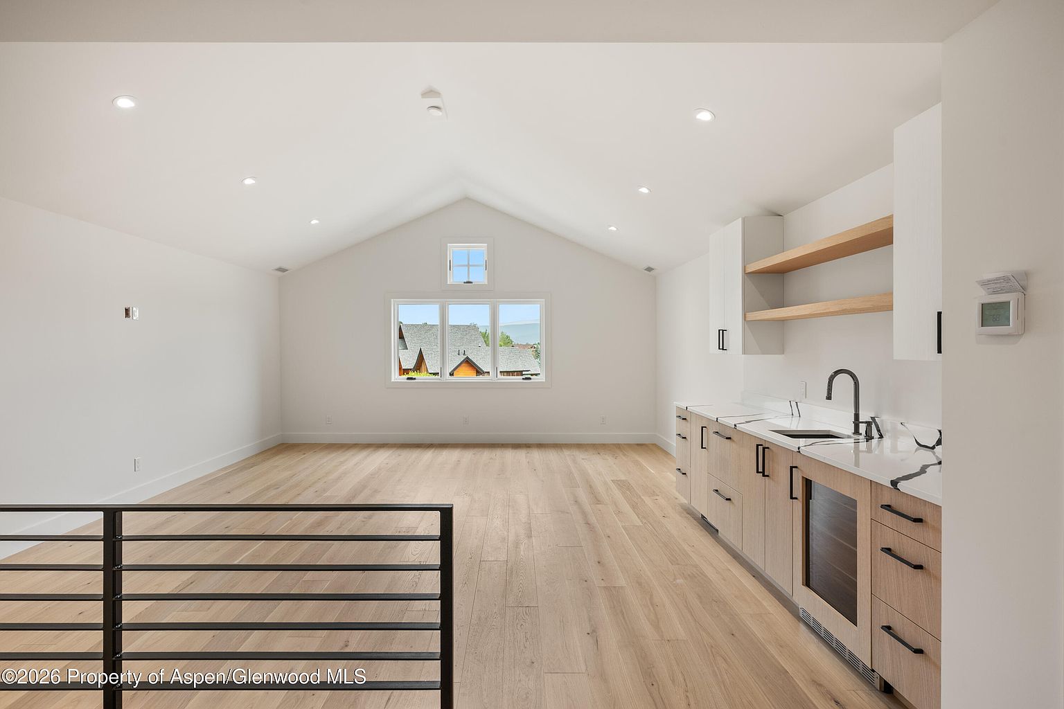 This bright, open-concept space features a kitchenette with light wood cabinetry, a built-in wine cooler, and a sink with a modern faucet. Floating shelves provide open storage above the counter, while the room is anchored by light-toned wood flooring and a vaulted ceiling with recessed lighting. A large window at the far end offers a view of the outdoors, creating an airy and versatile living area.