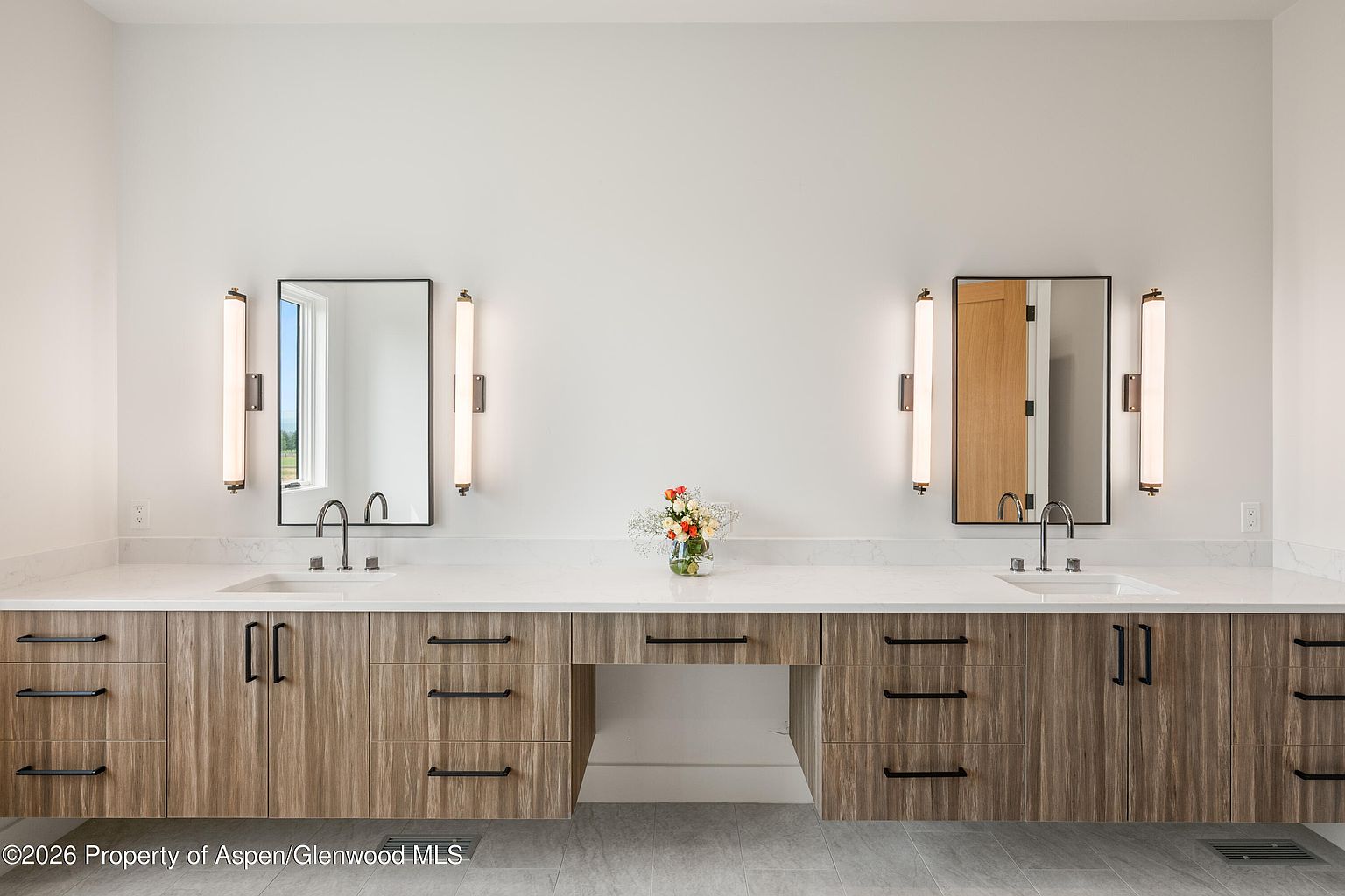 This primary bathroom features a long, dual-sink vanity with light wood-toned cabinetry and a clean, white countertop. The space is symmetrically designed with two rectangular mirrors flanked by vertical wall sconces, creating a balanced and modern aesthetic. A central open knee-space provides a vanity area, while a small vase of flowers adds a touch of color to the neutral, contemporary design.