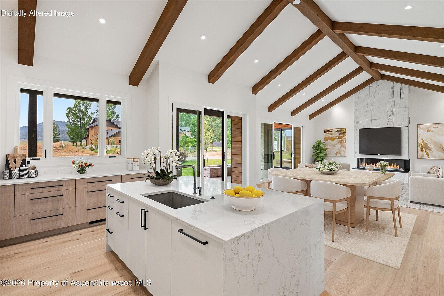 This open-concept kitchen and dining area features a bright, airy aesthetic with vaulted ceilings accented by exposed wooden beams. A large central island with a stone countertop and integrated sink serves as the focal point, complemented by light wood cabinetry and a dining table set for six. Large glass doors provide a seamless transition to the outdoors, while a tall, modern fireplace with a stone-clad chimney breast anchors the adjacent living space.