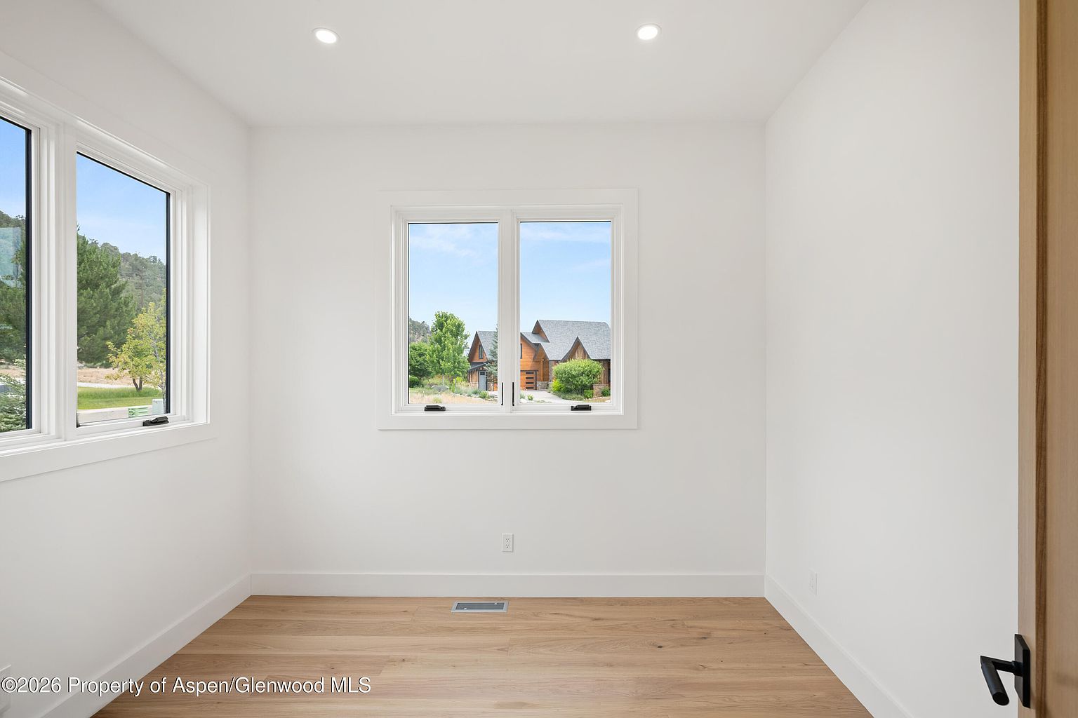 This bright, minimalist room features light wood flooring and crisp white walls, creating a clean and versatile space suitable for an office or study. Two large windows provide ample natural light and offer a pleasant view of the surrounding landscape and neighboring homes. The room is finished with recessed ceiling lighting and a central floor vent, presenting a neutral canvas ready for personalization.
