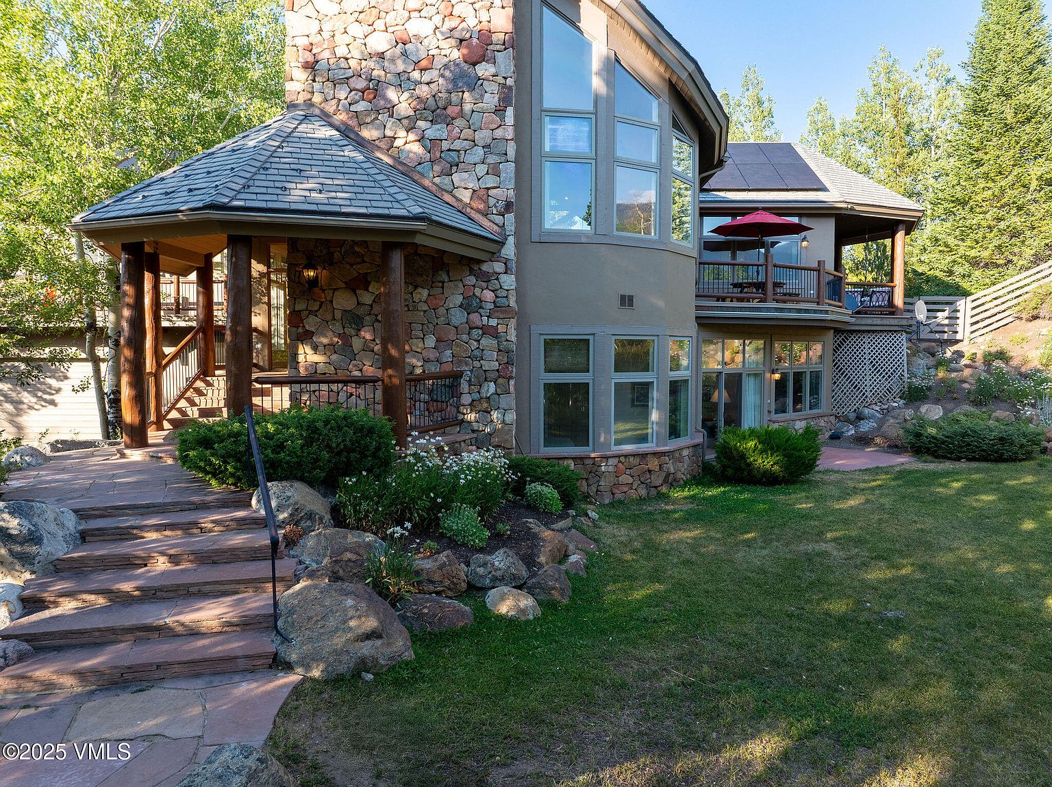 This is a front exterior view of a multi-story stone and stucco house. The house features a stone turret-like structure on the left, a covered porch, and a second-story balcony with a red umbrella. The landscaping includes a stone pathway, rock features, and well-maintained lawn, creating a welcoming and upscale impression.