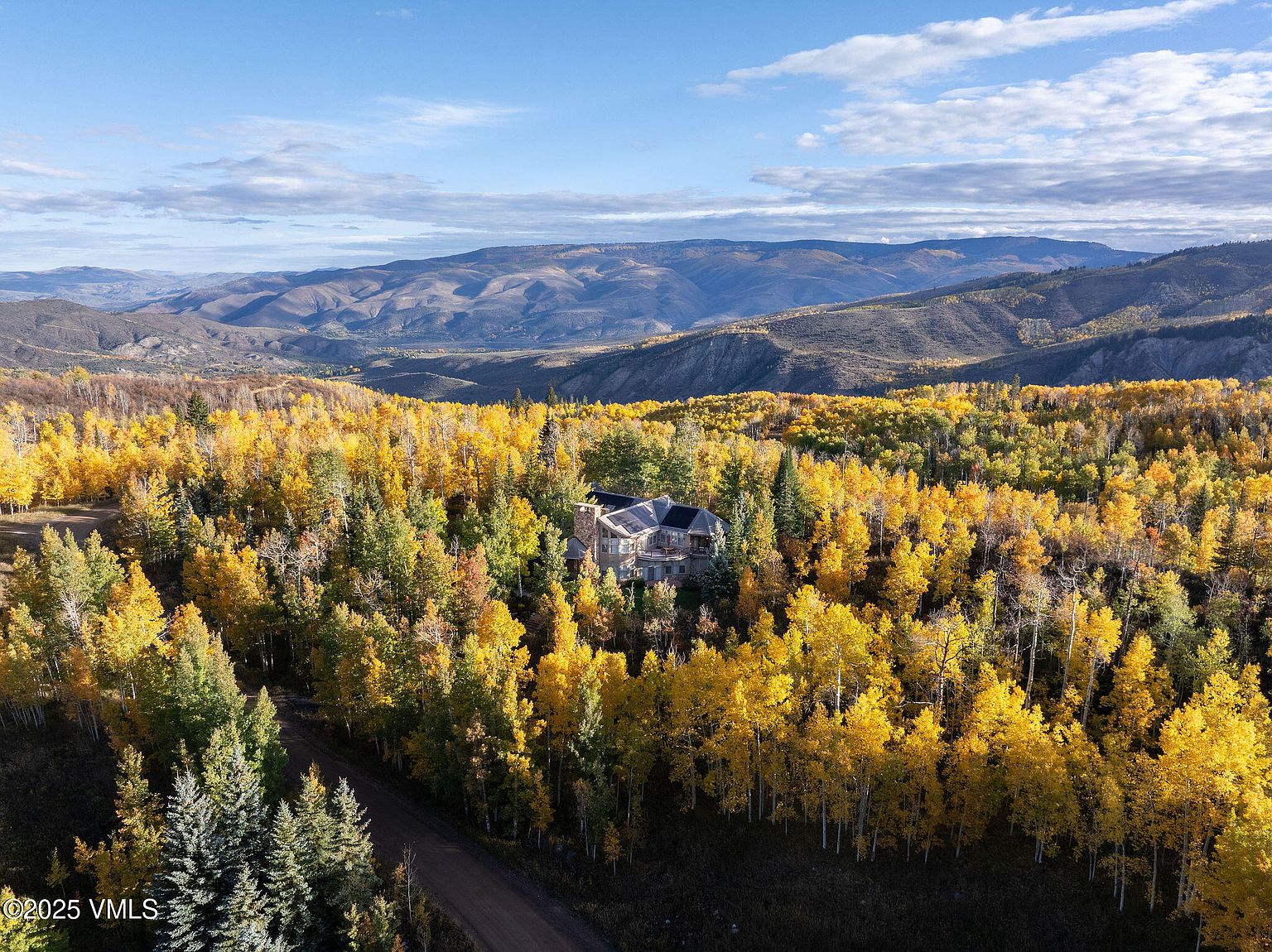 This aerial view showcases a luxurious home nestled within a vibrant forest displaying autumn colors. The house features a dark roof and is surrounded by a mix of evergreen and deciduous trees. In the background, rolling hills and a blue sky with scattered clouds create a picturesque landscape, emphasizing the property's secluded and scenic location.