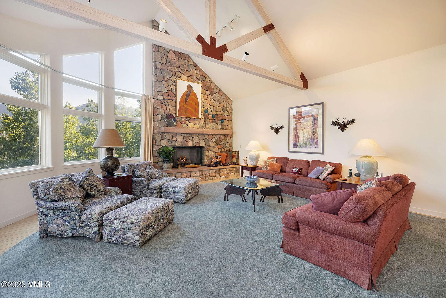 This is a spacious living room featuring a stone fireplace as a focal point, complemented by exposed wooden beams on the vaulted ceiling. The room is furnished with comfortable seating arrangements, including sofas and armchairs, set upon a neutral-toned carpet. Large windows provide ample natural light, enhancing the warm and inviting atmosphere of the space.