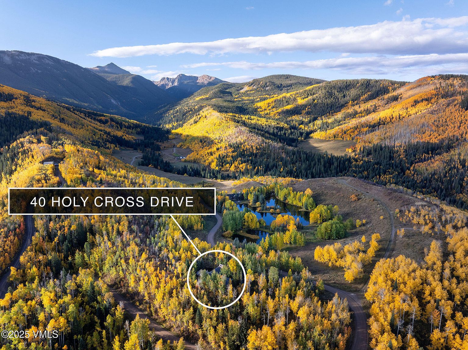 This aerial view showcases a property nestled within a vibrant autumn landscape, featuring rolling hills covered in golden and green trees. A winding dirt road leads to a secluded house, partially hidden among the trees, with a small pond nearby reflecting the colorful foliage. The scene is framed by distant mountains under a clear blue sky, creating a sense of tranquility and natural beauty.