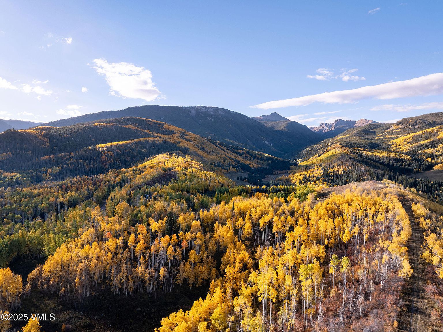 This aerial view showcases a vast landscape of rolling hills and mountains covered in a dense forest with vibrant yellow and green foliage, indicating the autumn season. The scene is bathed in sunlight under a clear blue sky with scattered clouds, creating a picturesque and serene environment. The perspective emphasizes the scale and natural beauty of the property, highlighting its potential for privacy and scenic views.
