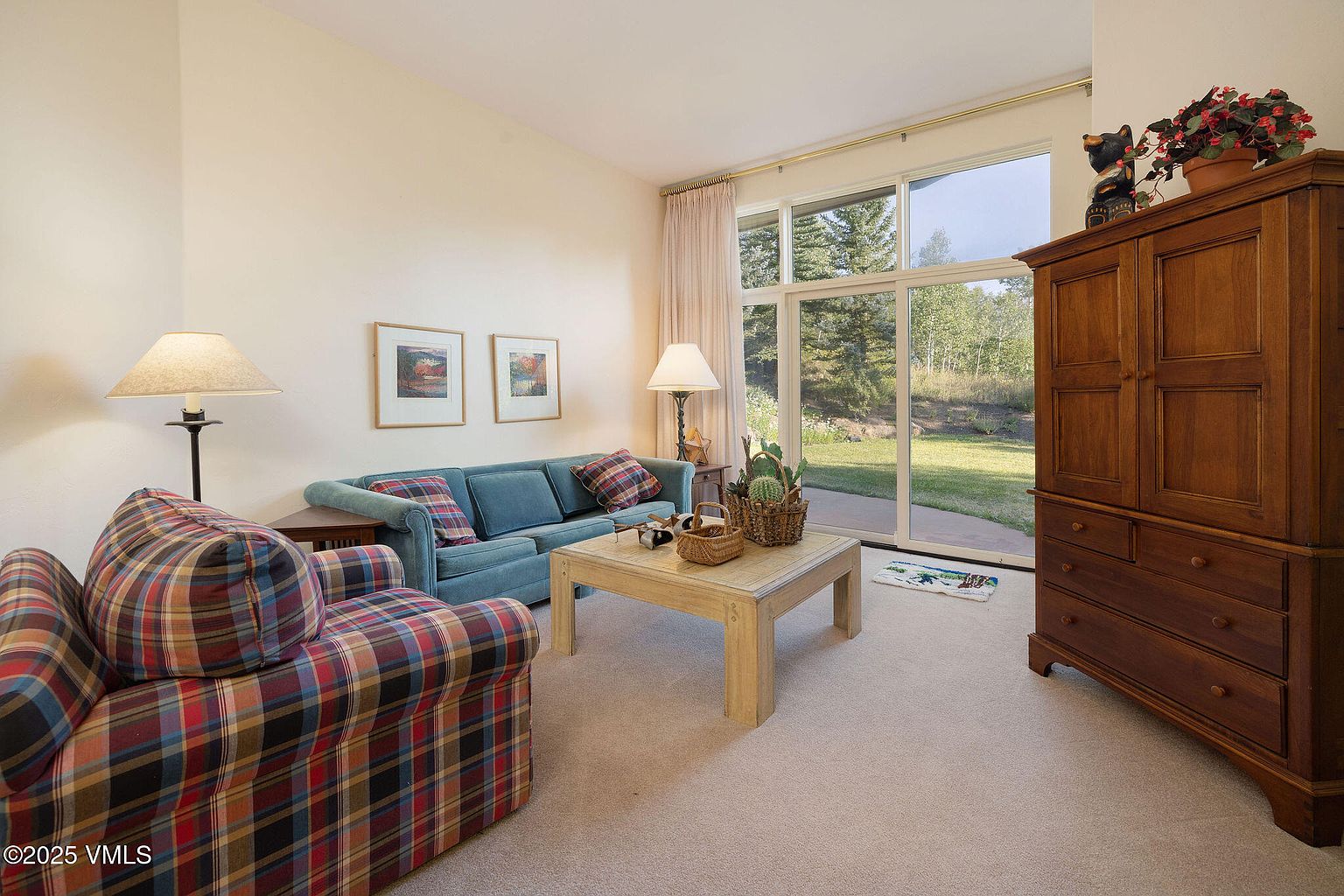 This is an interior shot of a living room featuring a blue sofa with plaid pillows, a plaid armchair, and a light wood coffee table. A large window provides natural light and a view of the outdoors, while a wooden cabinet stands against the wall. The room is carpeted and decorated with framed artwork and lamps, creating a cozy and inviting atmosphere.