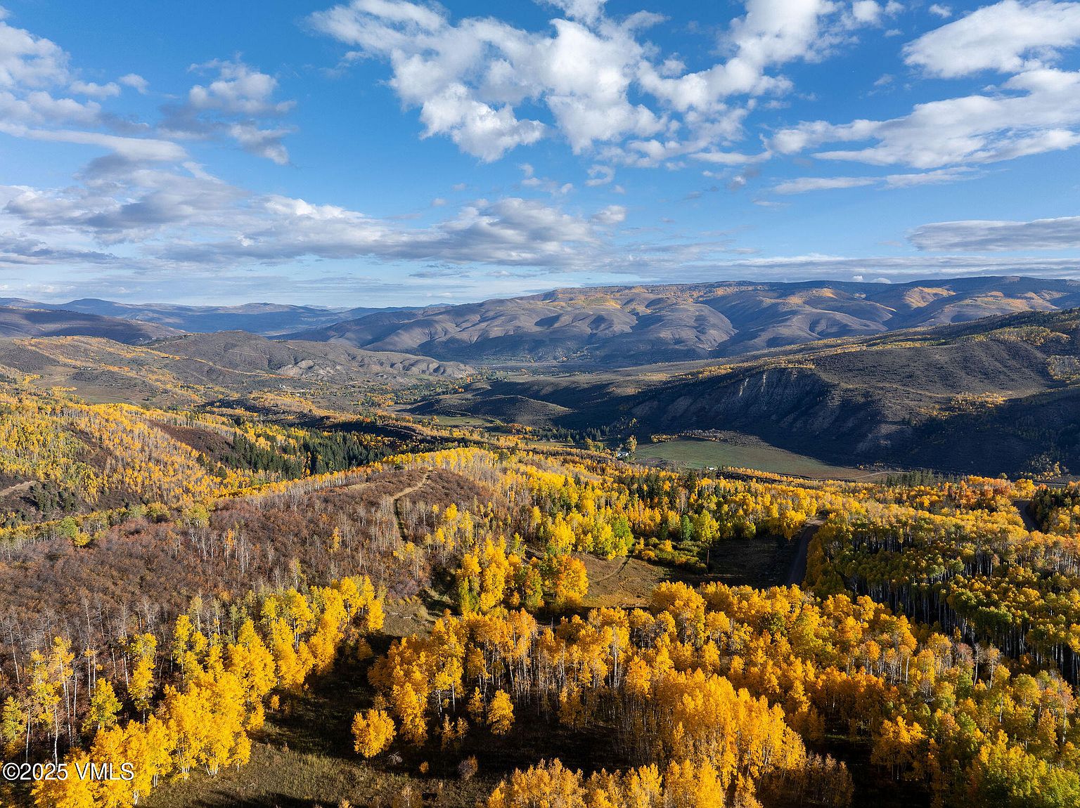 This aerial view showcases a vast landscape with rolling hills and mountains in the background, under a bright blue sky dotted with fluffy white clouds. The foreground is dominated by vibrant yellow aspen trees in their full autumn glory, creating a stunning contrast with the darker green patches of evergreen trees and the brown hillsides. The scene evokes a sense of tranquility and natural beauty, highlighting the property's access to expansive views and natural surroundings.