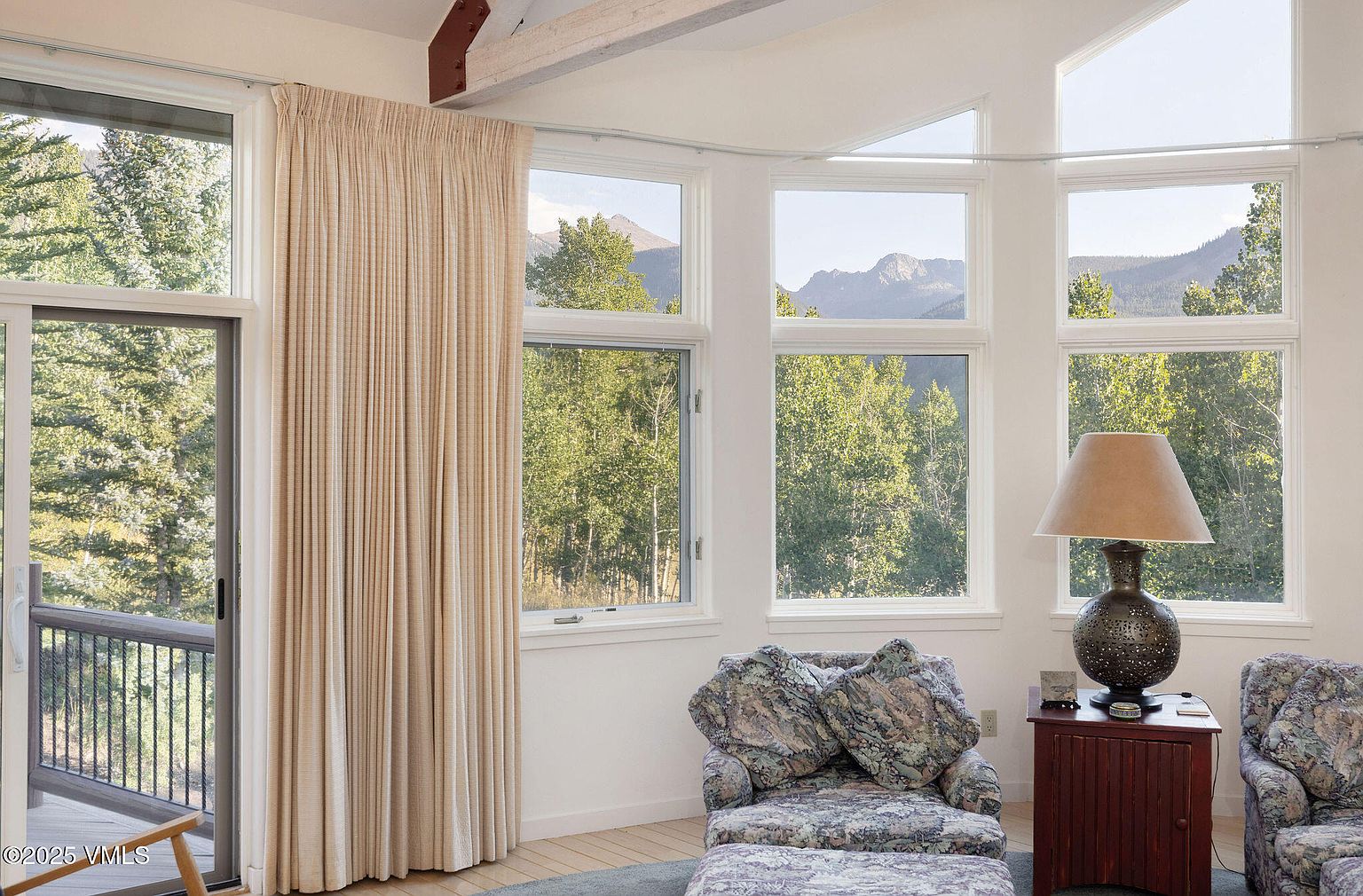 This is an interior shot of a living room featuring a seating area with patterned armchairs and a side table with a lamp. Large windows offer views of the surrounding trees and mountains, bringing natural light into the space. A curtain is drawn to the side of a sliding glass door that leads to a balcony.