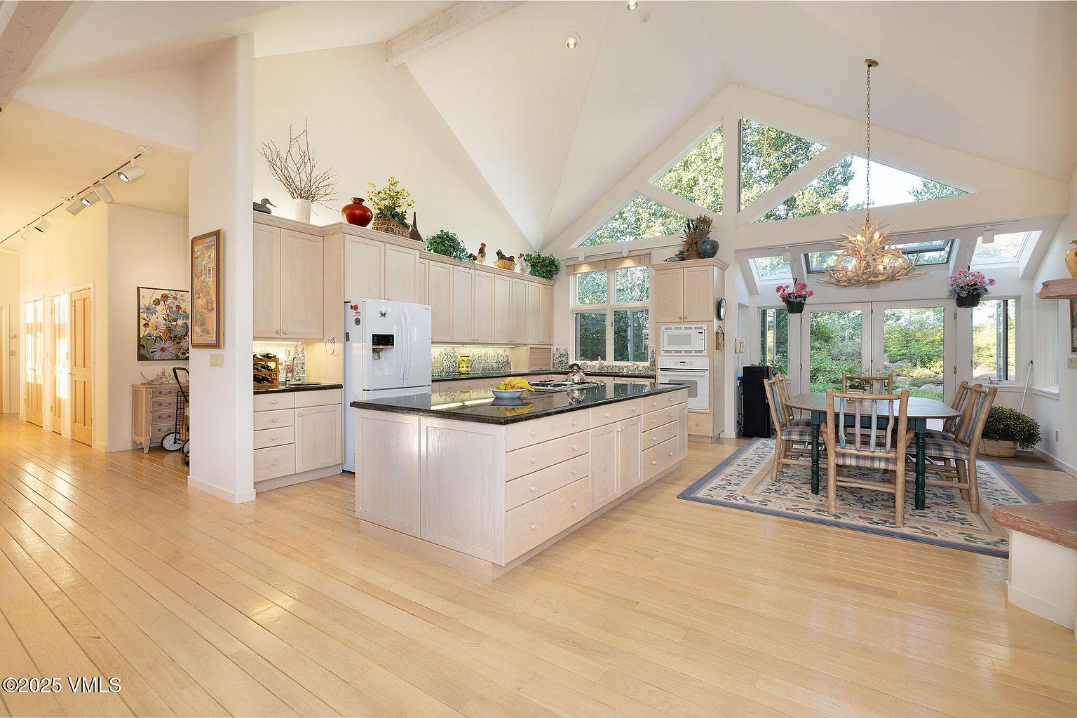 This is a bright and spacious kitchen featuring light wood cabinetry, a large center island with a black countertop, and a dining area with a table and chairs. The kitchen boasts a high vaulted ceiling with large windows that provide ample natural light and views of the outdoors. The overall impression is one of warmth, openness, and functionality, making it an inviting space for cooking and entertaining.