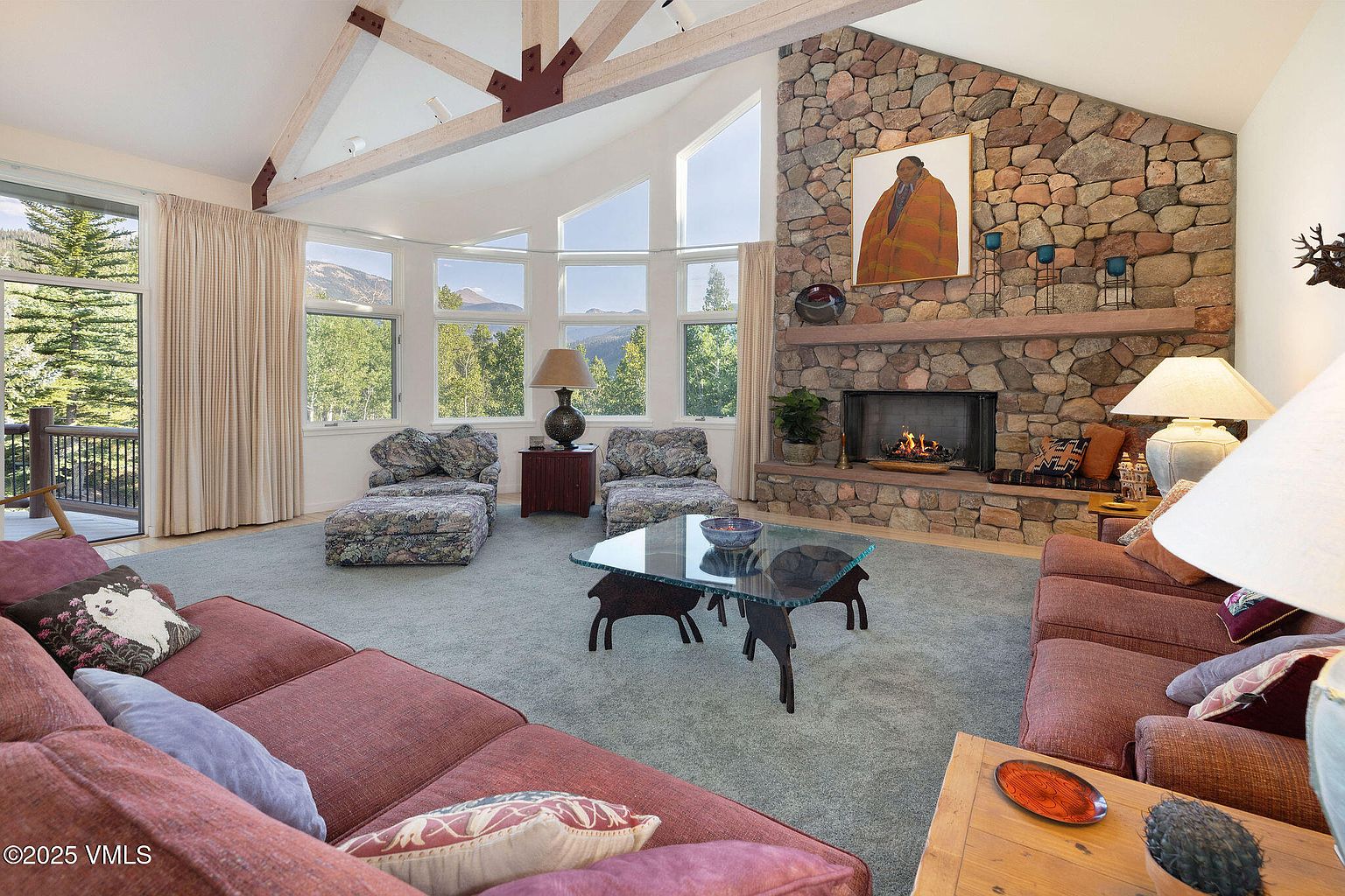 This is an interior shot of a living room featuring a stone fireplace, large windows with mountain views, and comfortable seating. The room has a rustic yet elegant feel, with exposed wooden beams and a neutral color palette. The layout is inviting and spacious, ideal for relaxation and entertaining.