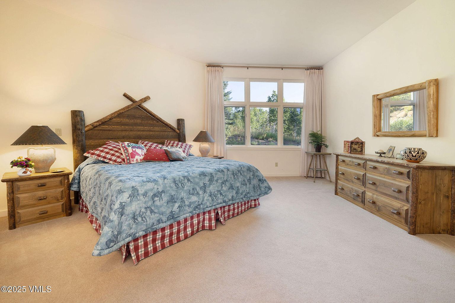 This is a cozy primary bedroom featuring a rustic wooden headboard and matching side tables and dresser. The bed is adorned with a blue patterned comforter and red plaid accents, creating a warm and inviting atmosphere. Natural light floods the room through a large window, enhancing the room's serene ambiance.