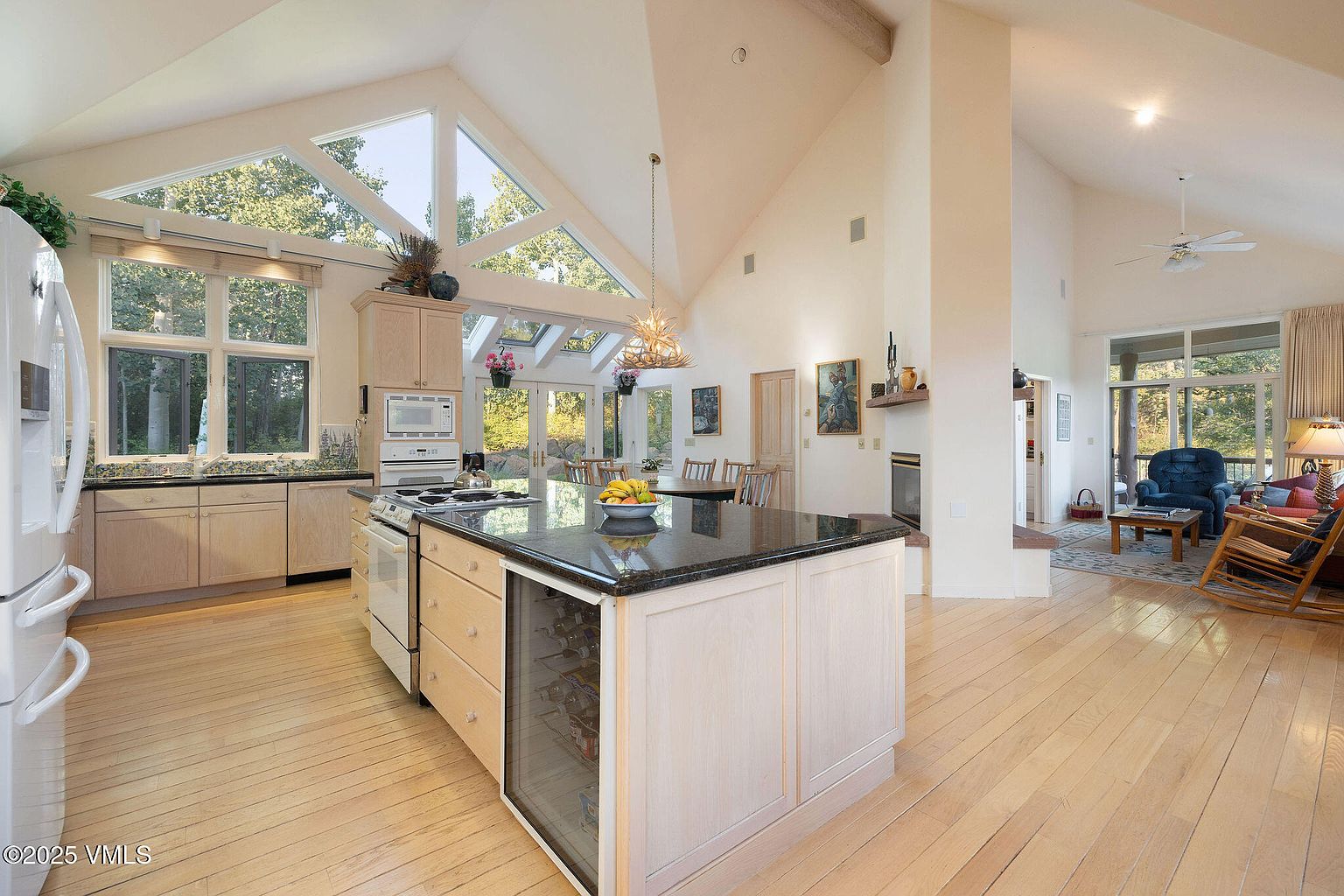 This is a bright and spacious kitchen featuring light wood cabinetry, a dark granite countertop island with a built-in wine cooler, and stainless steel appliances. The room boasts high vaulted ceilings with large windows that provide ample natural light and views of the surrounding greenery. The open floor plan seamlessly connects the kitchen to the living area, creating an inviting and airy atmosphere.