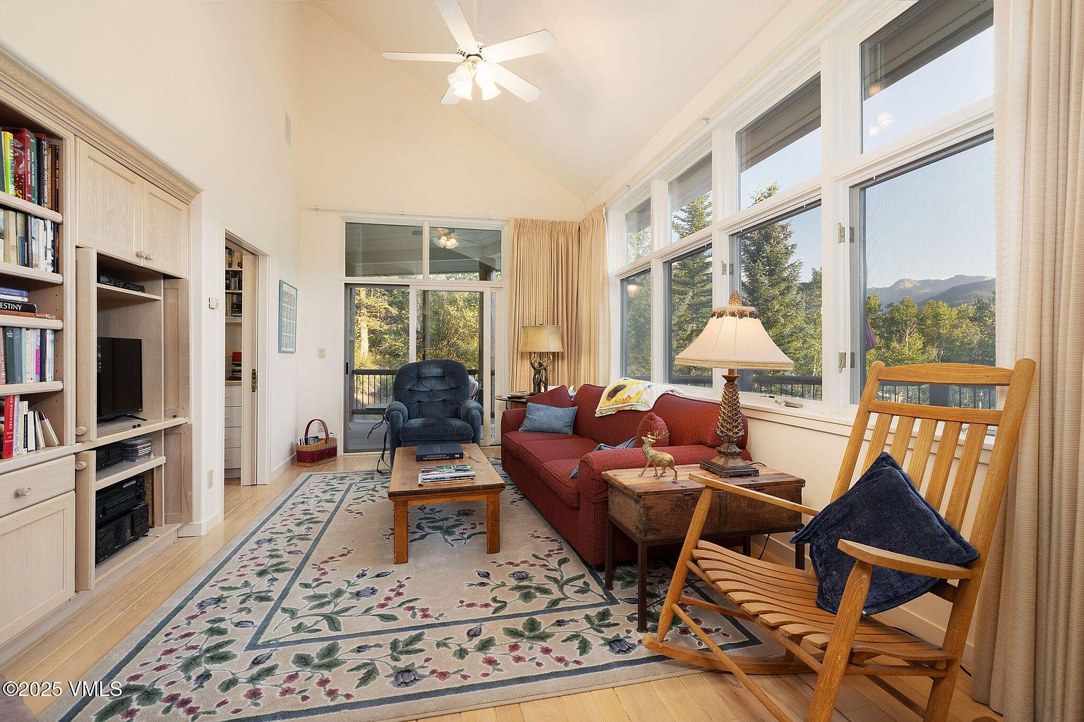 This is a cozy living room featuring a red sofa, a wooden rocking chair with a blue pillow, and a floral rug. Large windows provide ample natural light and views of the surrounding trees and mountains. A built-in cabinet with a TV and bookshelves adds functionality and charm to the space.