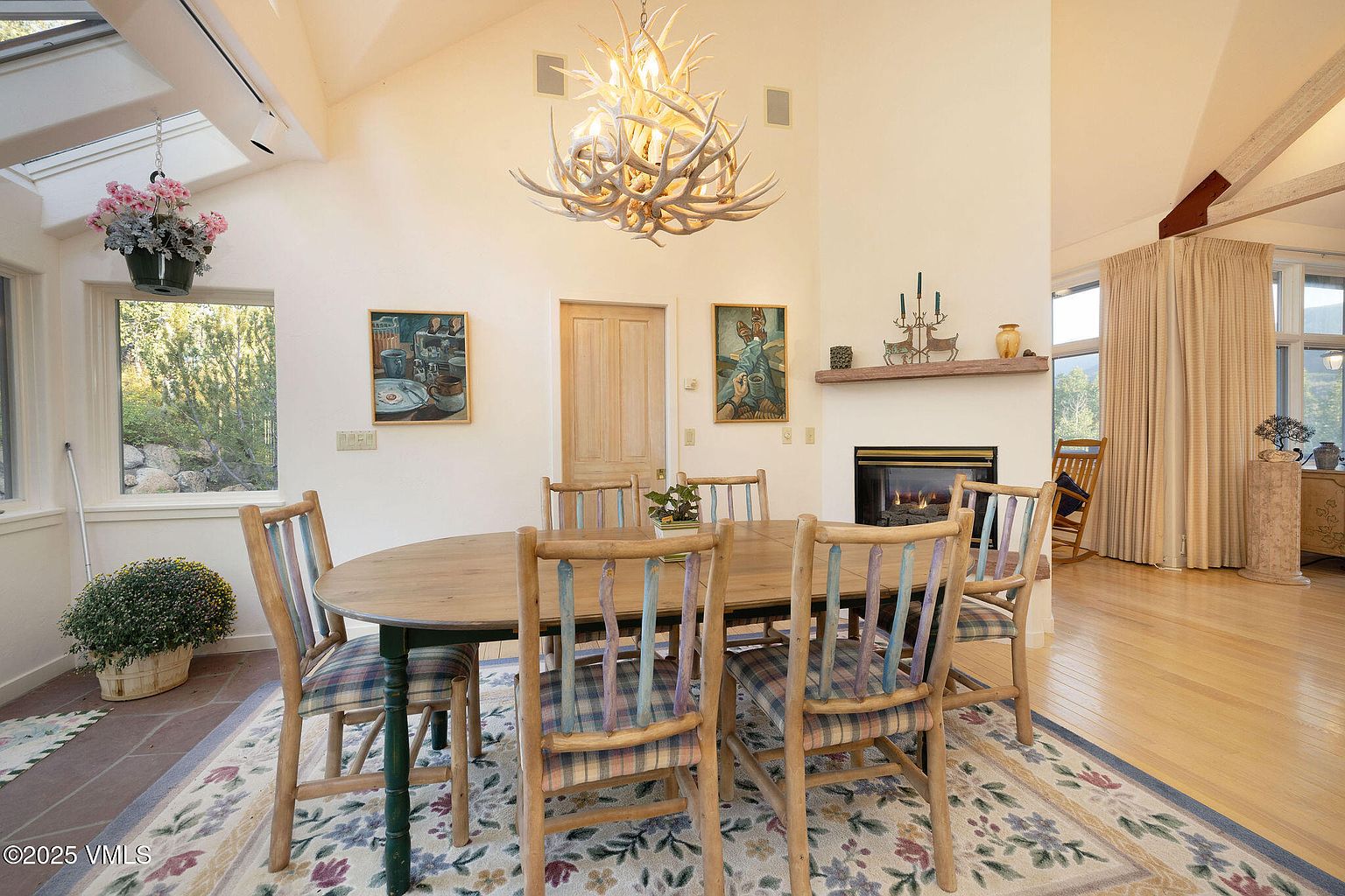 This is an interior shot of a dining room featuring a wooden oval table surrounded by six chairs with plaid cushions. Above the table hangs a unique antler chandelier, and artwork adorns the walls. A fireplace is visible in the background, adding warmth to the room. The room has a cozy and rustic feel.