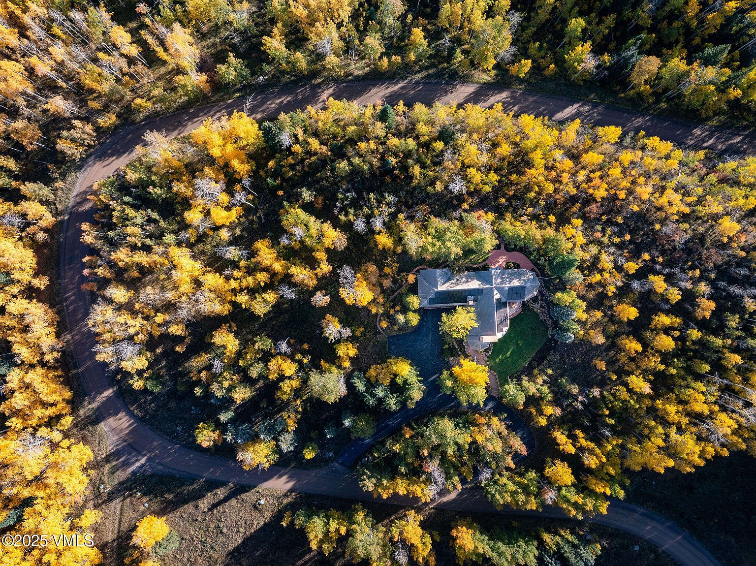 This aerial shot showcases a luxurious home nestled within a vibrant forest of autumn foliage. A winding road encircles the property, providing access and enhancing the sense of seclusion. The house features a complex roofline and is surrounded by mature trees, creating a picturesque and private setting.