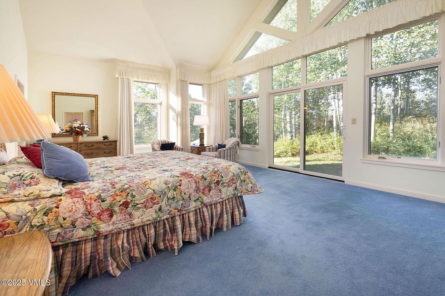 This is a bright and spacious primary bedroom featuring large windows that offer views of the surrounding greenery. The room is carpeted in blue and includes a bed with a floral comforter, a dresser with a mirror, and two armchairs. The high ceilings and natural light create an airy and inviting atmosphere.