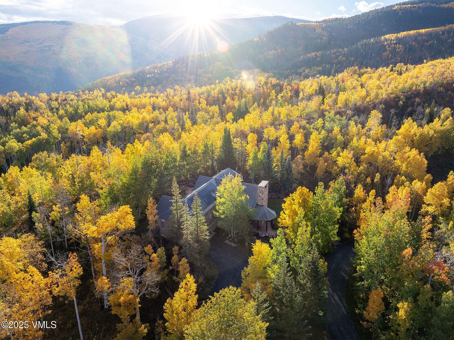This aerial shot showcases a beautiful home nestled within a vibrant forest in full autumn colors. The house features a stone chimney and a dark roof, blending harmoniously with the surrounding trees. The sun shines brightly, creating a warm and inviting atmosphere, highlighting the property's secluded and picturesque setting.