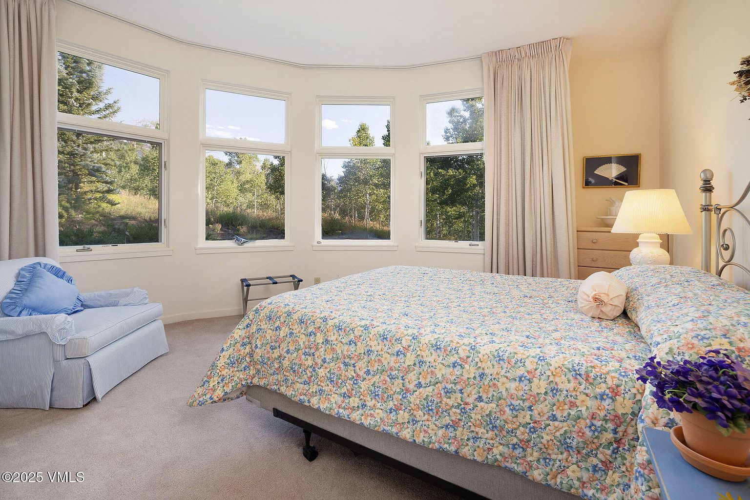 This is a cozy primary bedroom featuring a floral patterned bedspread, a comfortable armchair, and multiple windows offering natural light and views of the surrounding trees. The room is decorated in soft, neutral tones, creating a relaxing and inviting atmosphere. A bedside table with a lamp and a dresser provide functional storage.