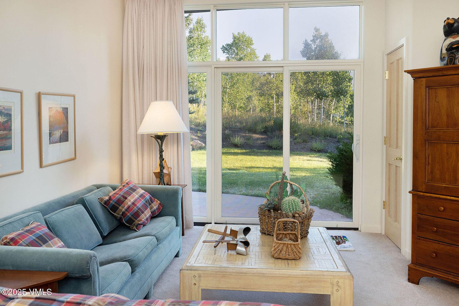 This is an interior shot of a living room featuring a light blue sofa with plaid pillows, a light-colored coffee table with decorative items, and a large window offering a view of a green lawn and trees. The room is well-lit, with a lamp providing additional illumination, and the overall style is comfortable and inviting, suggesting a relaxed living space.