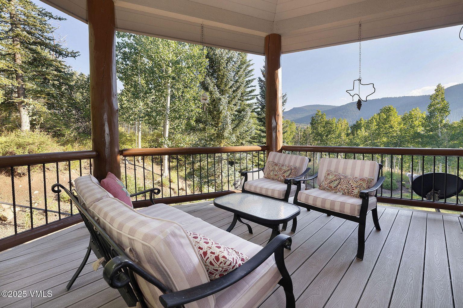 This image showcases a charming outdoor patio or deck area, furnished with comfortable seating including a sofa and two chairs arranged around a glass-topped table. The deck is constructed with light-colored wood planks, and a black metal railing provides safety and a touch of elegance. The backdrop features lush greenery and distant mountains, creating a serene and inviting atmosphere.