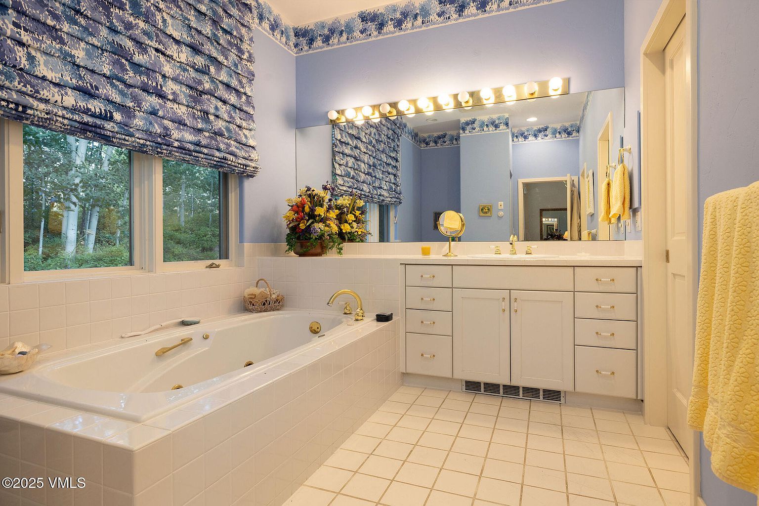 This is a well-lit primary bathroom featuring a large soaking tub with gold fixtures, a double vanity with white cabinetry, and a large mirror with vanity lighting. The walls are painted a soft blue, complemented by a floral wallpaper border and matching window treatment. The overall impression is clean, spacious, and classic.