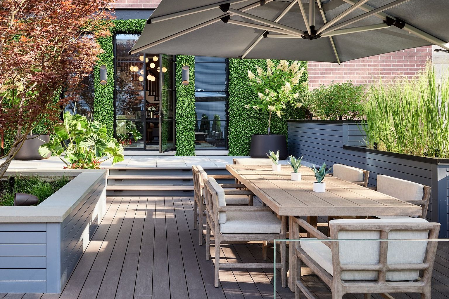 This sophisticated outdoor terrace features a spacious wooden deck furnished with a large dining table and cushioned chairs, perfect for al fresco entertaining. The space is beautifully landscaped with vertical green walls, a mature tree, and built-in planters, all shaded by a large, modern cantilever umbrella. The seamless transition from the deck to the interior through glass-paned doors creates an inviting, high-end atmosphere ideal for luxury living.