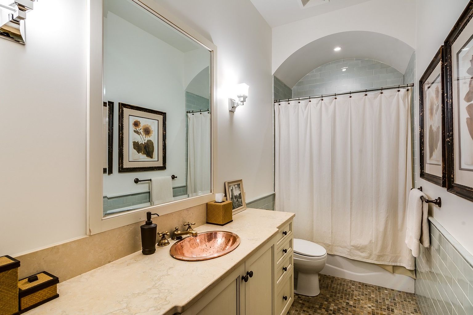 This elegant bathroom features a unique copper vessel sink set into a light-toned stone countertop, complemented by warm-toned cabinetry and a large wall-mounted mirror. The space includes a shower-tub combination with a classic white curtain and a soft, arched alcove design, creating a cozy and sophisticated atmosphere. The combination of light walls, subway tile accents, and warm metallic fixtures gives the room a refined, transitional aesthetic.