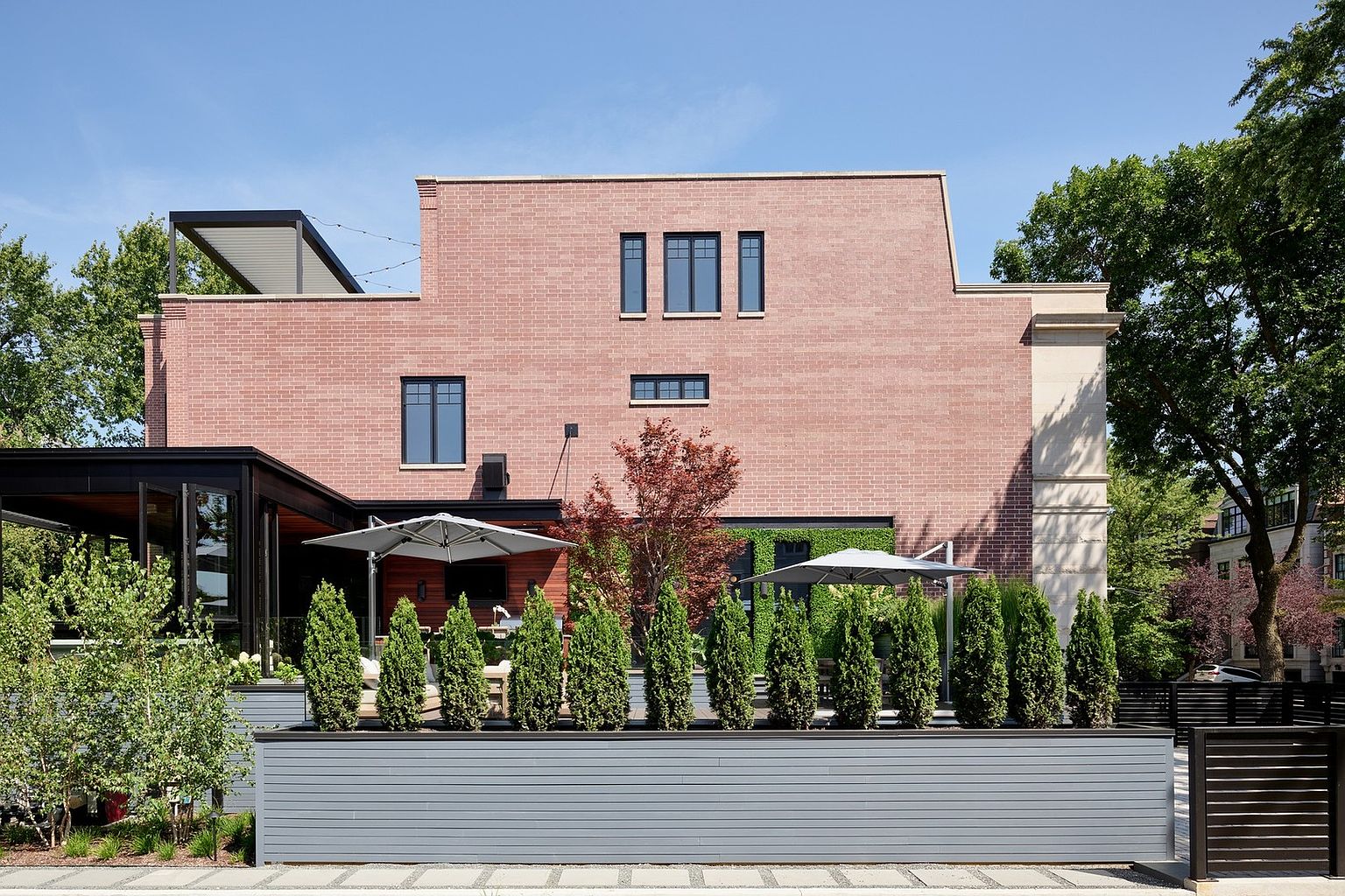 This image showcases the rear exterior of a modern, multi-story brick residence featuring a sophisticated patio area with a sleek gray privacy fence and a row of manicured evergreen shrubs. The space includes a covered outdoor living area with a dark metal frame, comfortable seating, and two large umbrellas, creating an inviting atmosphere for relaxation or entertaining. The architectural design blends traditional brickwork with contemporary elements, set against a backdrop of lush green trees and a clear blue sky.