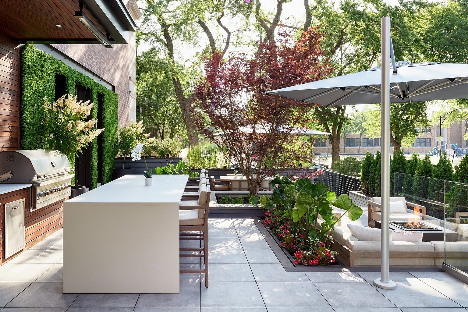 This sophisticated outdoor terrace features a sleek, modern design with a built-in stainless steel grill, a long minimalist dining island, and a cozy lounge area centered around a fire pit. The space is beautifully landscaped with a vertical green wall, lush potted plants, and a mature tree, all set against a backdrop of mature trees and a glass railing. The perspective is a wide, eye-level shot that captures the seamless blend of luxury outdoor living and urban greenery.