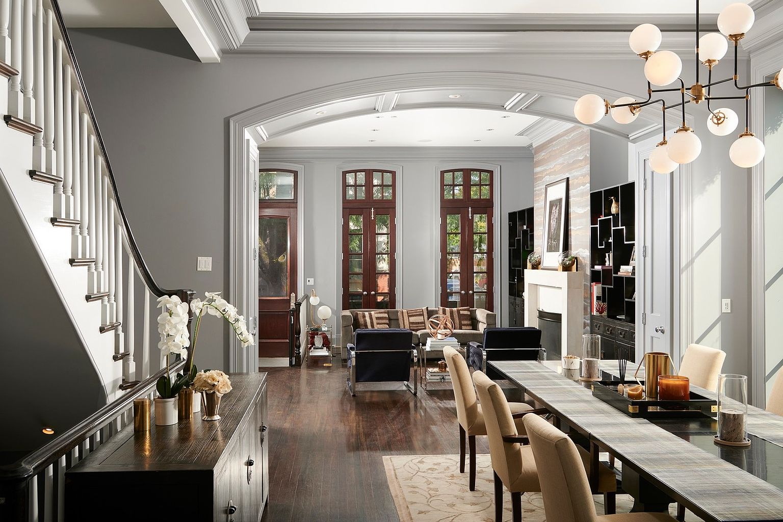 This elegant, open-concept space seamlessly transitions from a formal dining area in the foreground to a sophisticated living room beyond a grand arched entryway. The room features rich dark hardwood floors, a modern multi-globe chandelier, and a long dining table with upholstered chairs, all set against a backdrop of neutral gray walls and classic architectural molding. The perspective captures the depth of the home, highlighting the high-end finishes and the inviting, well-lit atmosphere.