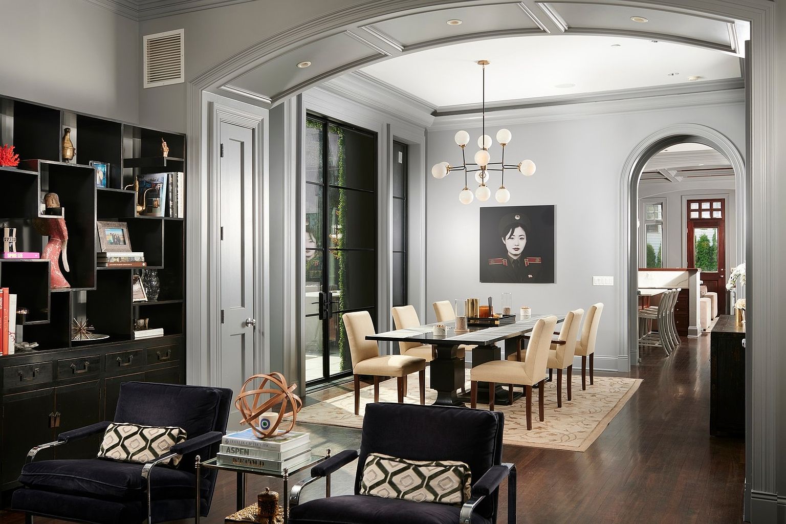 This elegant dining room features a sophisticated blend of modern and classic design, anchored by a dark wood table surrounded by cream-colored upholstered chairs. The space is illuminated by a striking contemporary globe chandelier and accented by a bold portrait on the wall, while a large black shelving unit and dark velvet armchairs in the foreground add depth and texture. The room flows seamlessly into an adjacent space through a graceful arched doorway, highlighting the home's refined architectural details and open, airy layout.