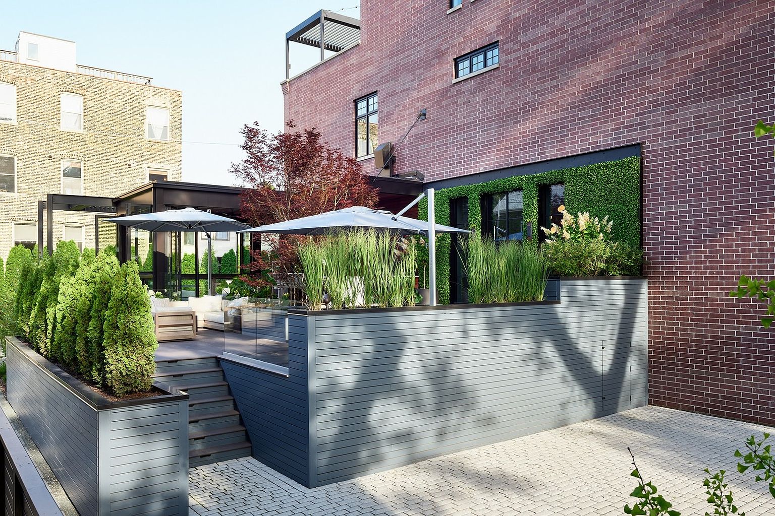 This sophisticated urban outdoor space features a multi-level deck with modern grey horizontal slat siding and integrated planters filled with lush greenery. The area includes a comfortable seating arrangement shaded by umbrellas, set against a backdrop of a classic red brick building with a vertical green wall. The perspective captures a clean, contemporary design that seamlessly blends architectural structure with natural elements, creating an inviting and private retreat.