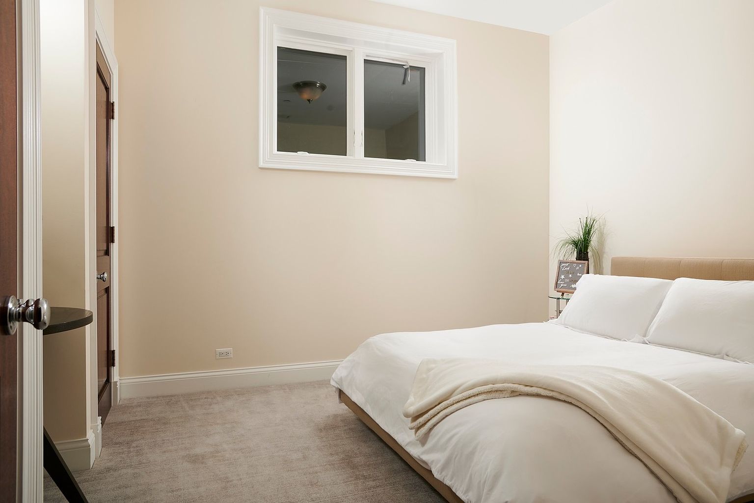 This cozy, minimalist bedroom features a neutral color palette with soft beige walls and plush grey carpeting. A comfortable bed with crisp white linens and a textured throw blanket serves as the focal point, positioned beneath a simple window that allows for natural light. The room is accented by a small side table with a decorative sign and a potted plant, creating a serene and inviting atmosphere.