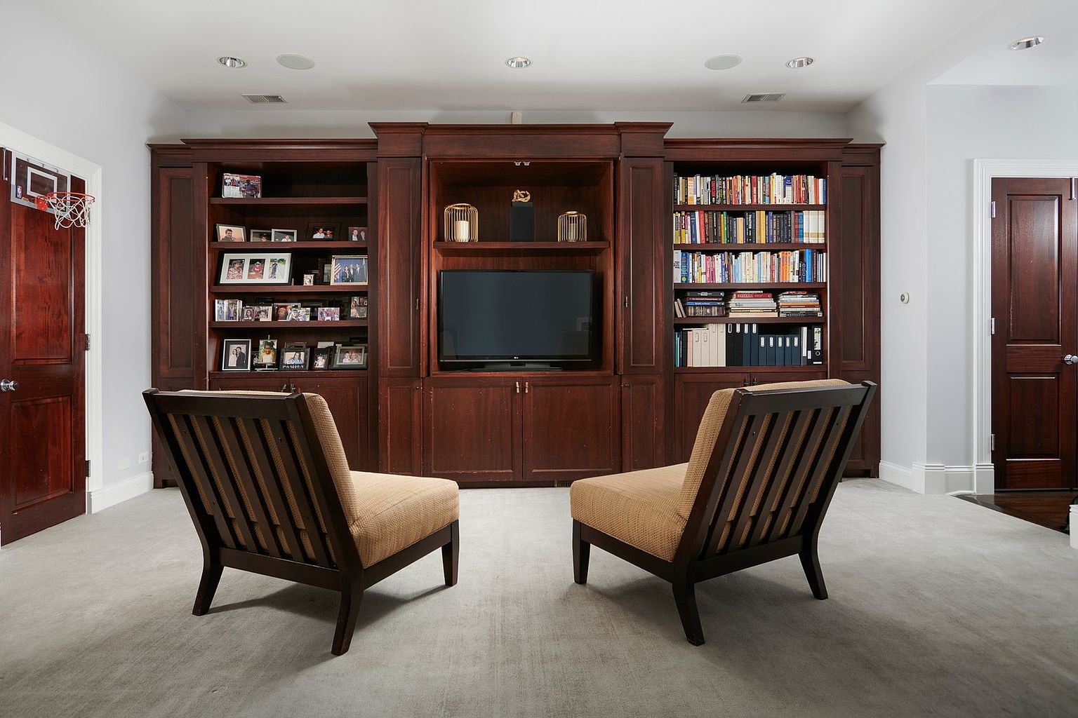 This sophisticated home office or study features a large, custom-built dark wood wall unit that serves as both a bookshelf and a media center for the television. Two comfortable, tan-upholstered armchairs with matching dark wood frames are positioned in the foreground, creating a cozy seating area. The room is carpeted in a neutral grey, and the presence of a small basketball hoop on the door adds a playful, personalized touch to the otherwise formal space.