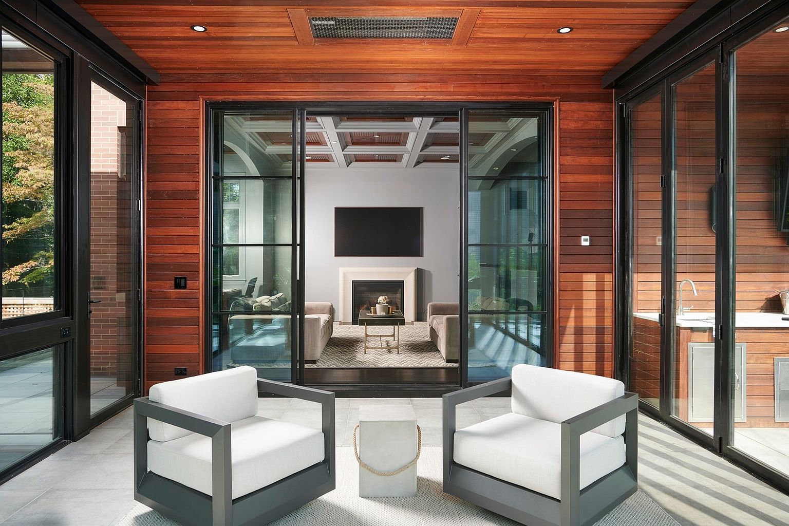 This sophisticated sunroom features warm wood-paneled walls and ceilings, creating a cozy yet modern atmosphere. Two contemporary white armchairs face a central stone side table, while large glass sliding doors provide a seamless transition to the interior living space beyond. The space is designed for relaxation, offering a bright and airy environment with a view into a refined, neutral-toned living room.