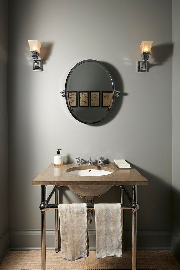 This elegant powder room features a classic pedestal-style vanity with a stone countertop and polished chrome legs. A circular mirror centered above the sink reflects framed botanical art, while matching wall sconces provide warm, symmetrical lighting. The neutral gray walls and tiled floor create a sophisticated, timeless aesthetic suitable for a high-end home.