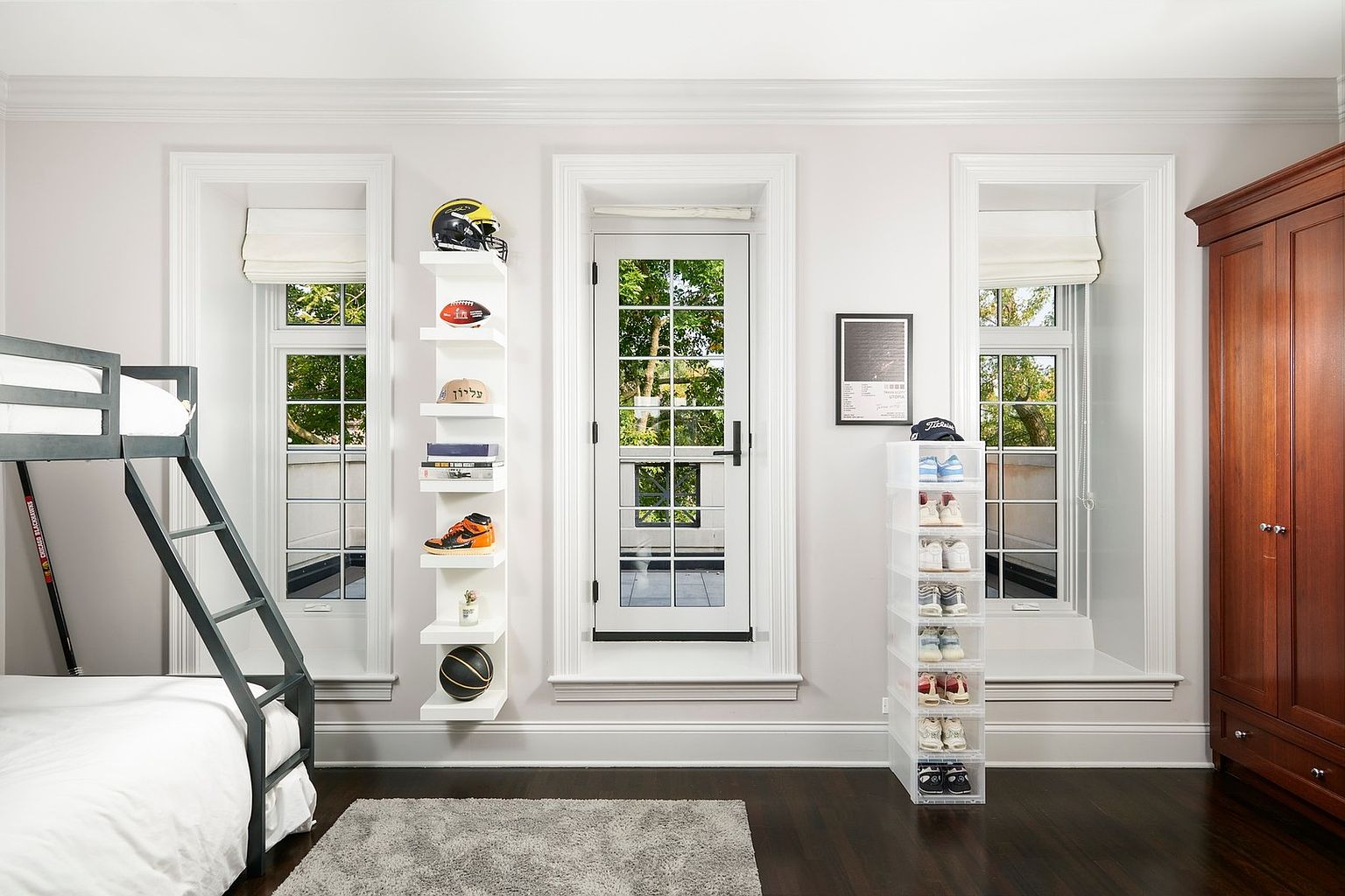 This well-organized bedroom features a modern bunk bed, a tall white display shelf showcasing sports memorabilia, and a clear acrylic shoe storage tower. The room is framed by three symmetrical windows and a central glass-paned door that opens onto a balcony, allowing for abundant natural light. A rich wooden wardrobe and dark hardwood floors provide a sophisticated contrast to the clean, contemporary decor.