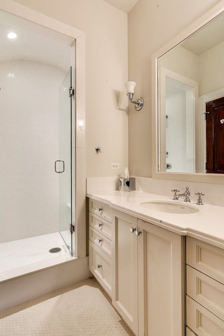 This bright and clean bathroom features a glass-enclosed walk-in shower with white penny-round tile flooring and walls. The vanity is equipped with a white countertop, a single undermount sink, and classic cabinetry, complemented by a large mirror and elegant wall sconce lighting. The overall aesthetic is neutral, polished, and inviting, making it a functional and stylish space.