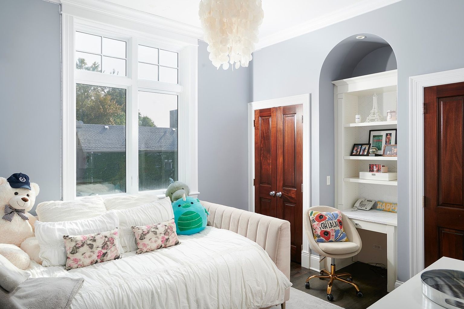 This charming bedroom features a soft, light blue color palette with a plush, cream-colored daybed adorned with decorative floral pillows and a large teddy bear. A built-in desk nook with shelving is tucked into an arched alcove, providing a cozy workspace, while a unique, textured chandelier adds a touch of elegance. The room is brightened by a large window and accented by rich, dark wood doors, creating a warm and inviting atmosphere.