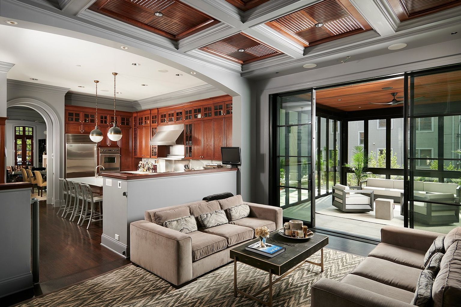 This sophisticated open-concept living space seamlessly blends a cozy, neutral-toned living room with a high-end gourmet kitchen featuring rich wood cabinetry and stainless steel appliances. A dramatic coffered ceiling with wood inlays adds architectural depth, while expansive floor-to-ceiling glass doors open to a covered outdoor patio, creating a fluid indoor-outdoor living experience. The perspective captures the elegant transition between the intimate seating area and the bright, airy kitchen, highlighting the home's luxurious and functional design.