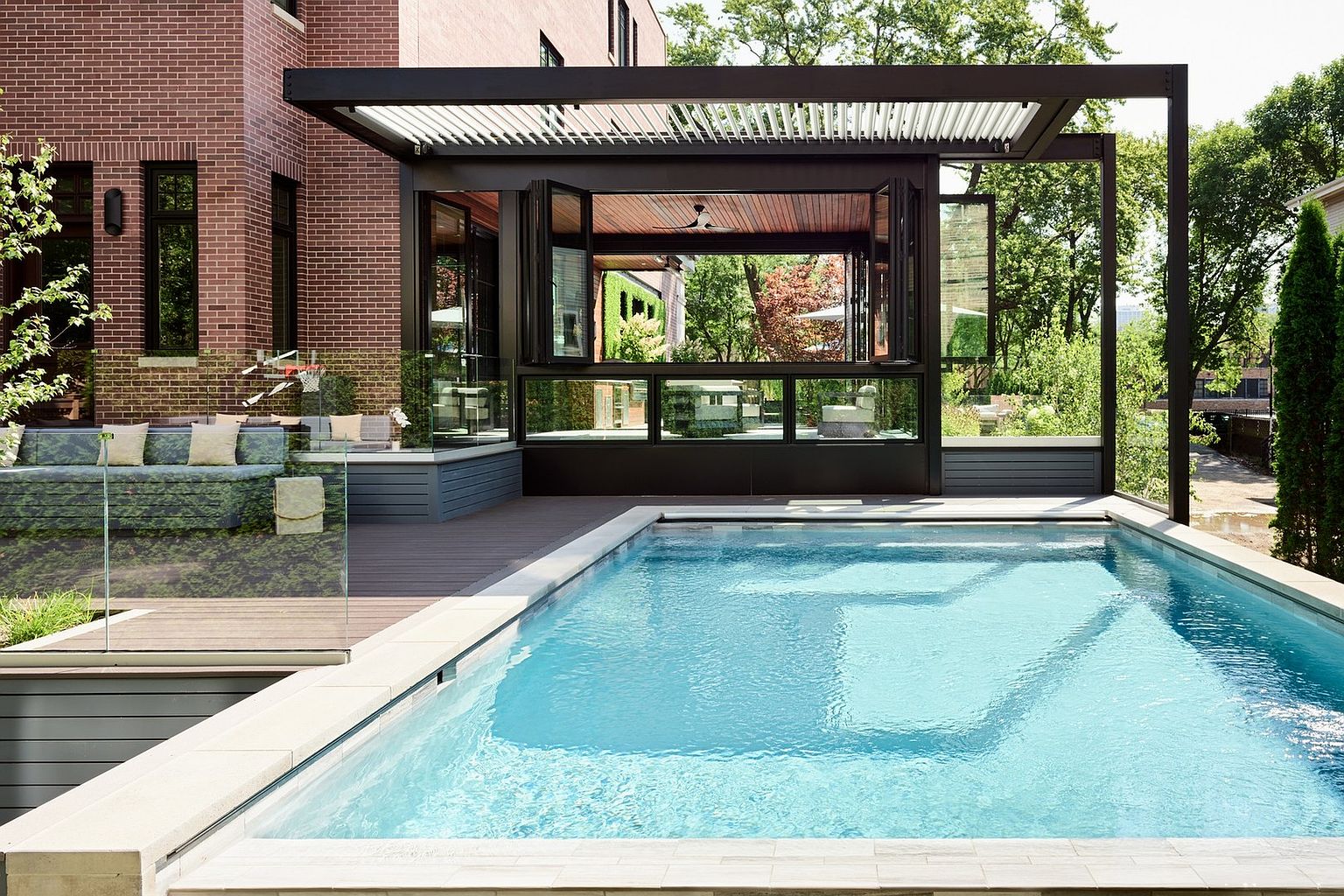 This sophisticated outdoor living space features a pristine, rectangular swimming pool set against a modern, multi-level deck and a brick-clad home. A striking black pergola covers a portion of the deck, which includes a seamless indoor-outdoor transition through large, folding glass doors that reveal a cozy interior lounge area. The combination of clean architectural lines, glass railings, and lush surrounding greenery creates a luxurious and inviting atmosphere perfect for high-end entertaining.
