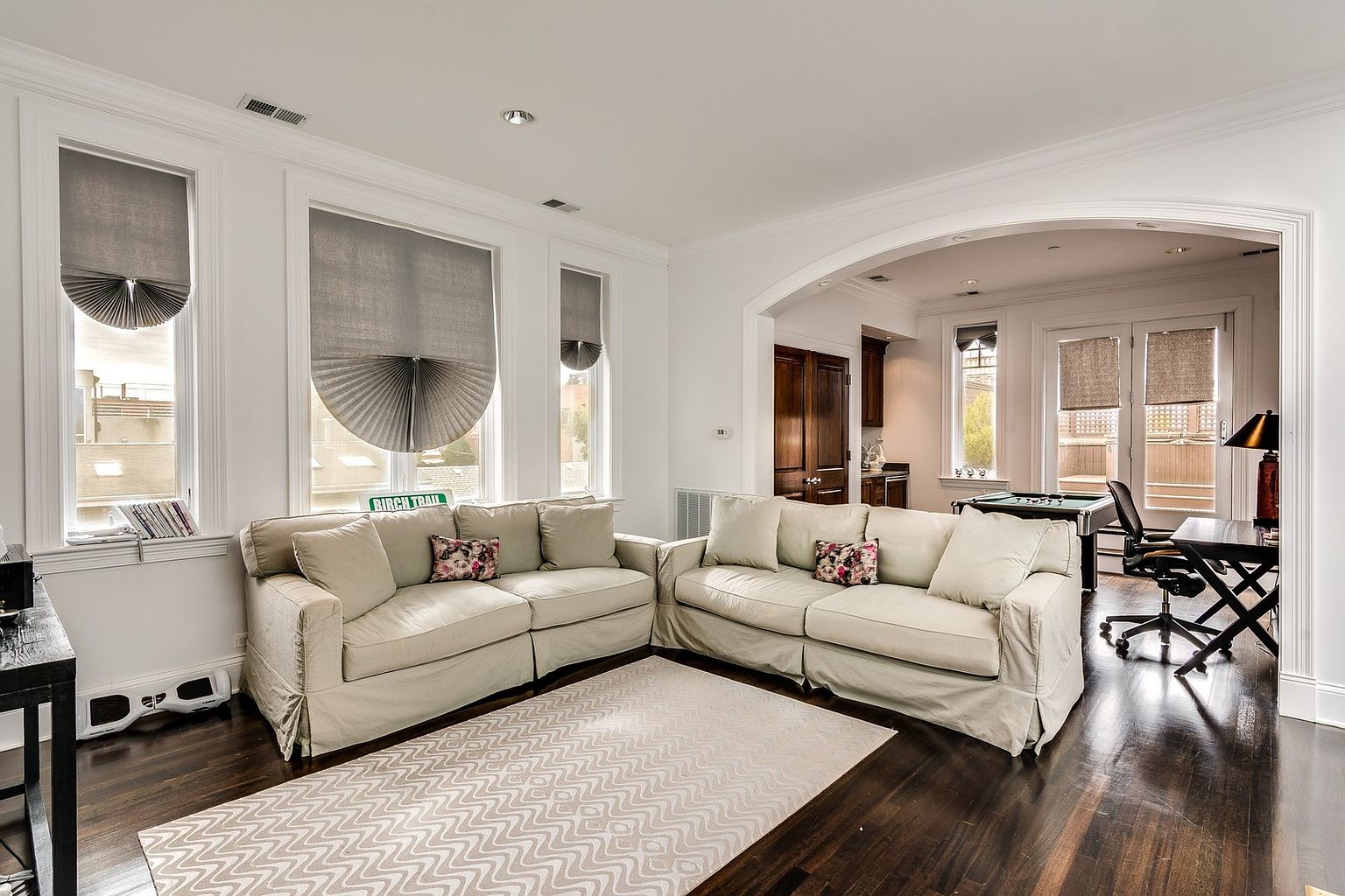 This bright and inviting living room features a large, cream-colored sectional sofa arranged on a patterned area rug over dark hardwood floors. The space is illuminated by tall windows with grey shades and flows seamlessly into an adjacent game room through a wide, arched entryway. The neutral color palette and clean architectural lines create a sophisticated and comfortable atmosphere perfect for relaxation or entertaining.