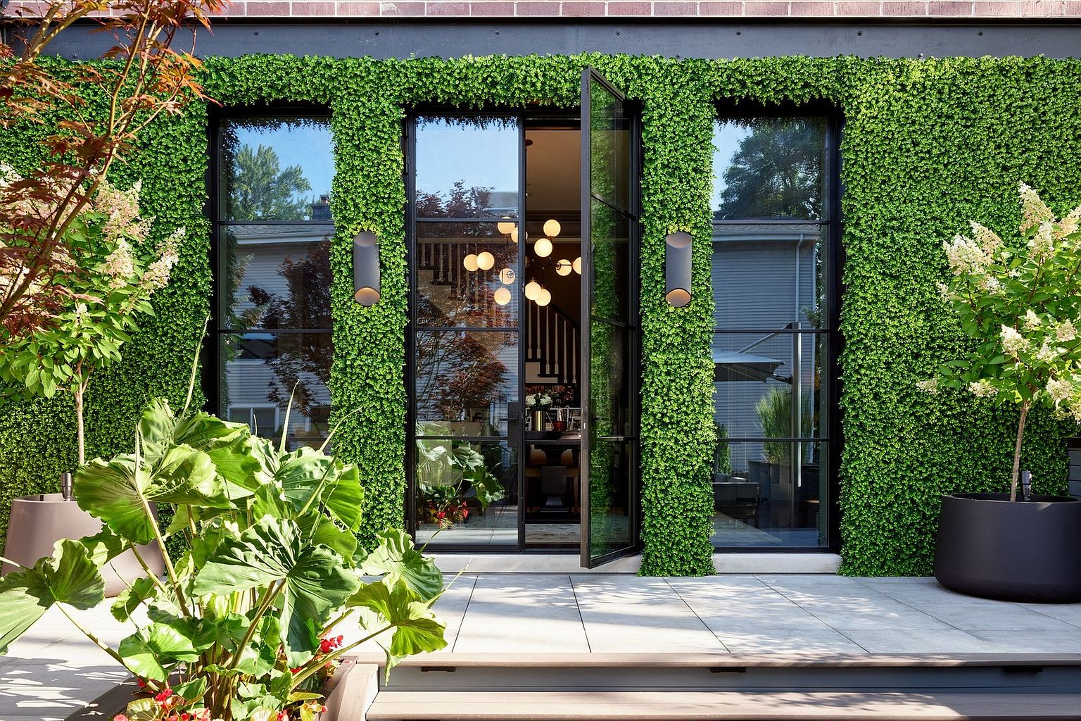 This sophisticated patio area features a striking living wall of lush greenery surrounding modern, black-framed glass doors that open into the home. The space is accented by large, contemporary planters and sleek wall-mounted lighting, creating a seamless blend between indoor and outdoor living. The clean lines and vibrant foliage provide a high-end, tranquil atmosphere perfect for entertaining.