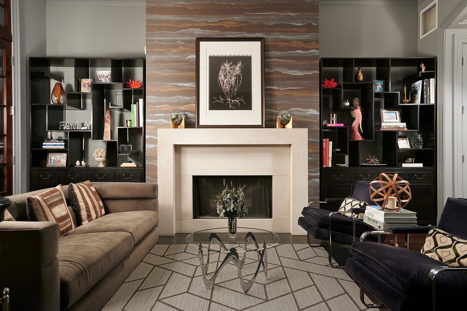 This sophisticated living room features a striking stone fireplace as its focal point, framed by custom floor-to-ceiling black shelving units. The space is anchored by a plush brown sofa and dark velvet armchairs, all arranged on a geometric-patterned area rug. A unique, modern glass coffee table sits in the center, while the textured, layered-look wallpaper adds depth and a contemporary, high-end aesthetic to the room.