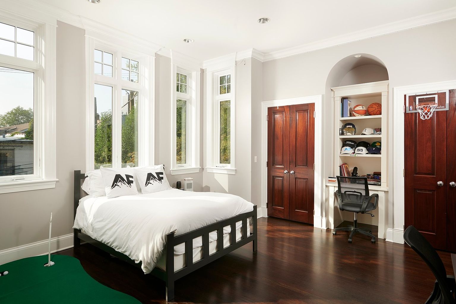 This spacious bedroom features dark hardwood floors, light grey walls, and a black metal bed frame adorned with white bedding. The room includes a built-in desk area with shelving, a closet with dark wood doors, and a basketball hoop mounted above the door, suggesting a versatile space for a child or teenager. Large windows allow ample natural light to fill the room, which also includes a small indoor putting green in the foreground.