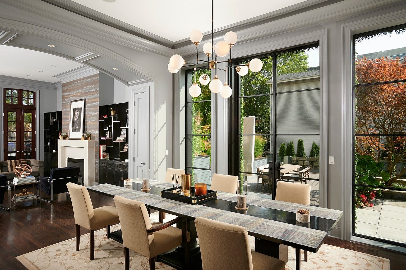 This elegant dining room features a long, modern table with a textured runner, surrounded by upholstered beige chairs, all illuminated by a striking contemporary globe chandelier. Large, floor-to-ceiling black-framed windows provide an expansive view of the outdoor patio and lush greenery, seamlessly blending indoor and outdoor living. The sophisticated space is anchored by a neutral area rug and flows into an adjacent living area with a fireplace, creating a cohesive and luxurious open-concept feel.