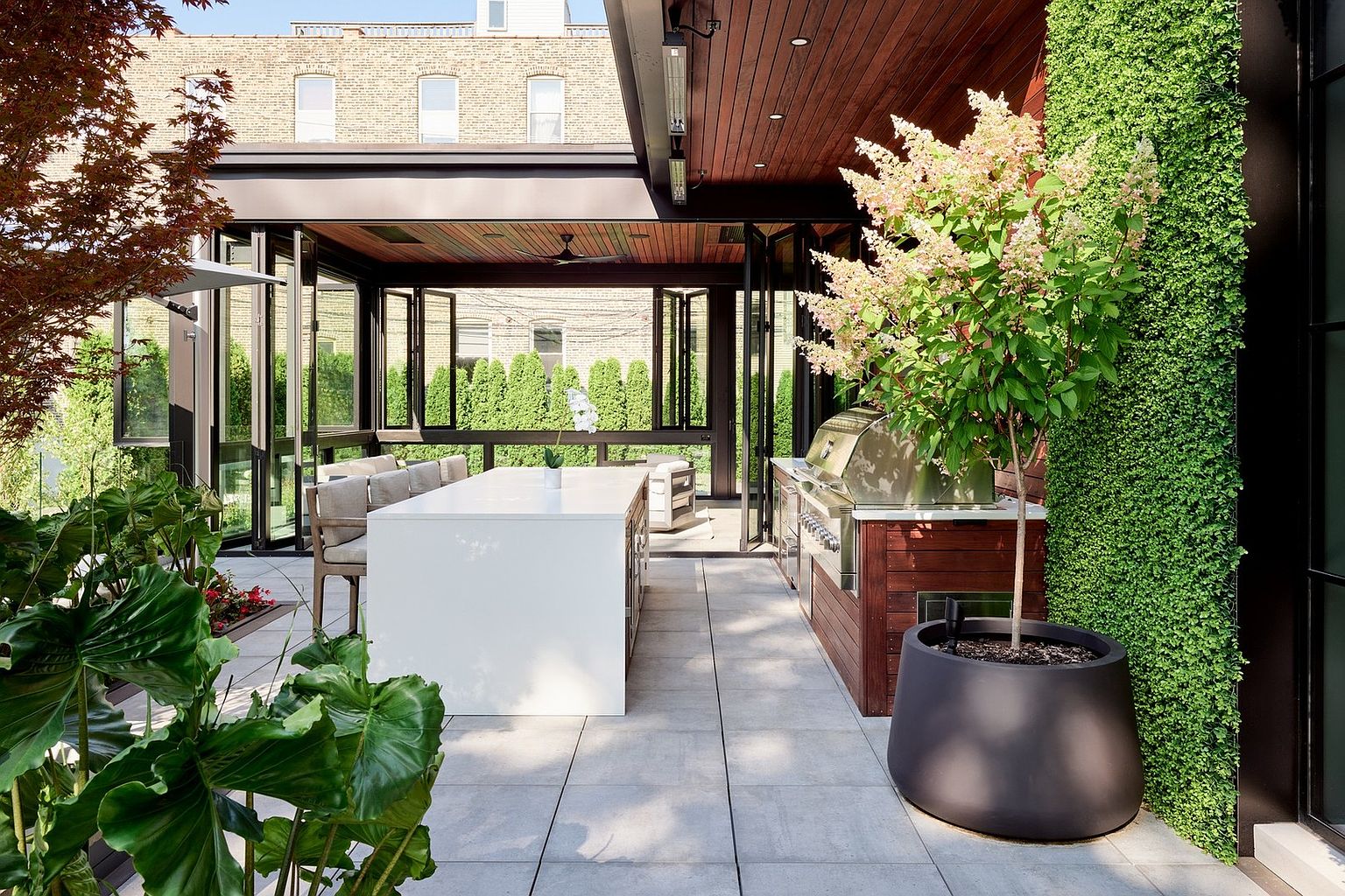 This sophisticated outdoor living space features a covered patio with a sleek white kitchen island and a built-in stainless steel grill. The area is framed by modern folding glass doors that open into an indoor space, complemented by a lush vertical green wall and a large potted tree. The clean lines, wood-paneled ceiling, and stone tile flooring create a luxurious, contemporary atmosphere perfect for high-end entertaining.
