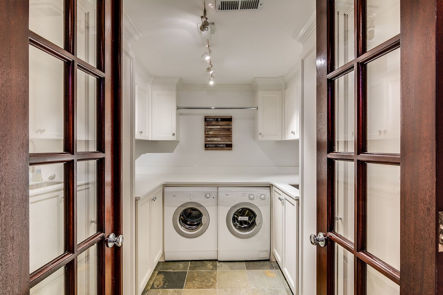 This laundry room is viewed through elegant, dark-stained French doors, creating a framed, inviting perspective. The space features clean white cabinetry, a sleek countertop, and a pair of modern front-loading washer and dryer units, all set against a durable slate-tiled floor. A decorative sign on the wall adds a touch of warmth to this functional and well-organized utility area.